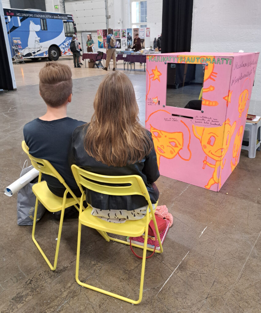 2 persons sitting in front of the Caricature Machine in Heslinki Comics festival