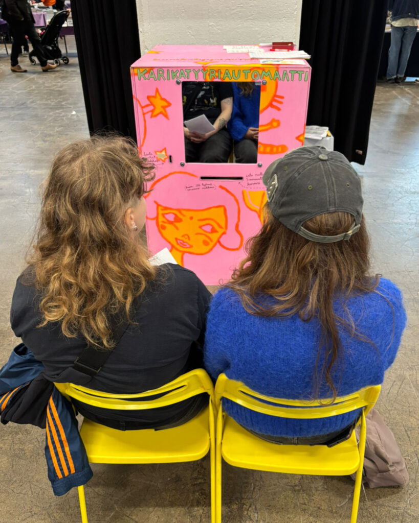 2 persons sitting in front of the Caricature Machine in Heslinki Comics festival